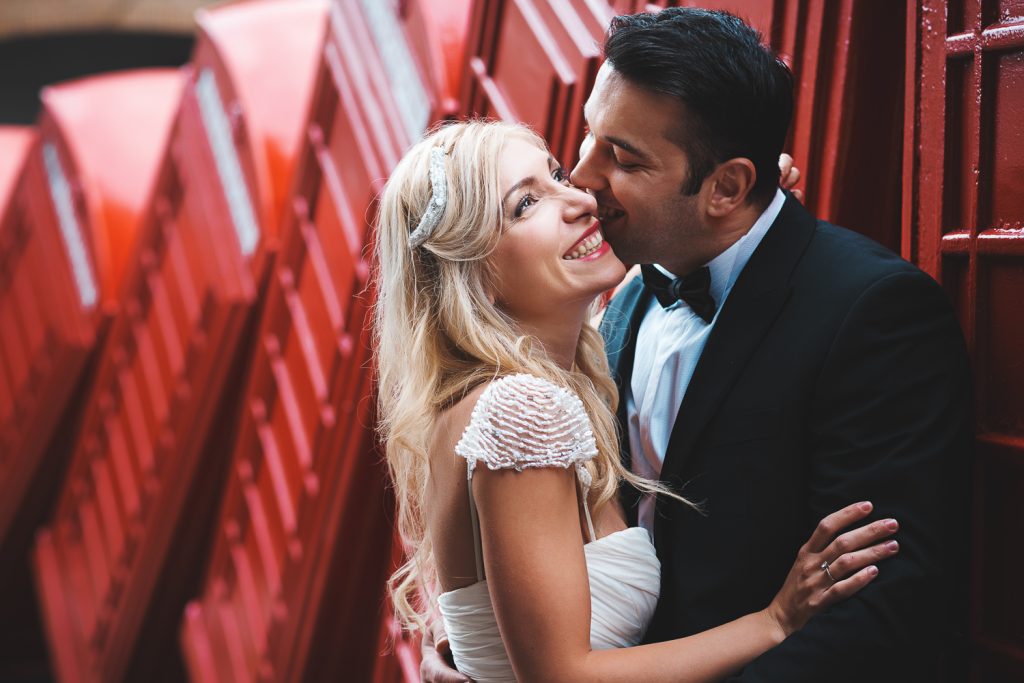 Happy bride smiling while kissed on cheek by groom during wedding photo shoot in London with red telephone boots in the background